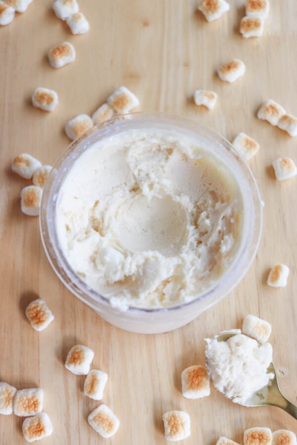 A container of marshmallow cream sits on a wooden surface, surrounded by small scattered marshmallows. A spoon with some cream on it rests nearby.