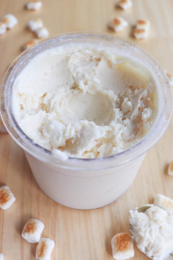 A plastic container filled with spreadable white butter, partially scooped out, sits on a wooden surface. Small toasted marshmallows are scattered around the container.