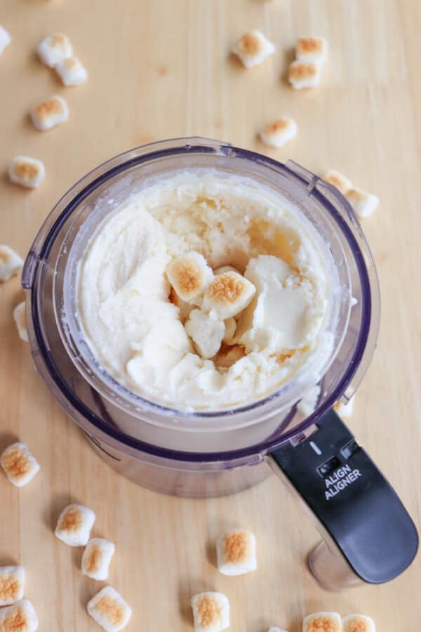 A food processor container filled with creamy mixture, topped with toasted mini marshmallows. More mini marshmallows are scattered on the wooden surface around the container.