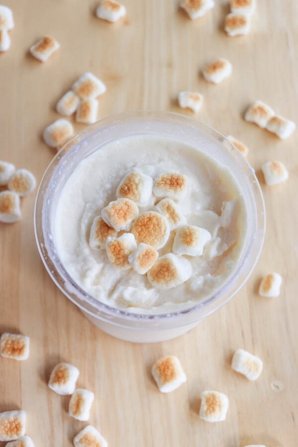 Vanilla ice cream topped with small toasted marshmallows in a glass container on a wooden surface. Scattered around the container are additional toasted marshmallows.