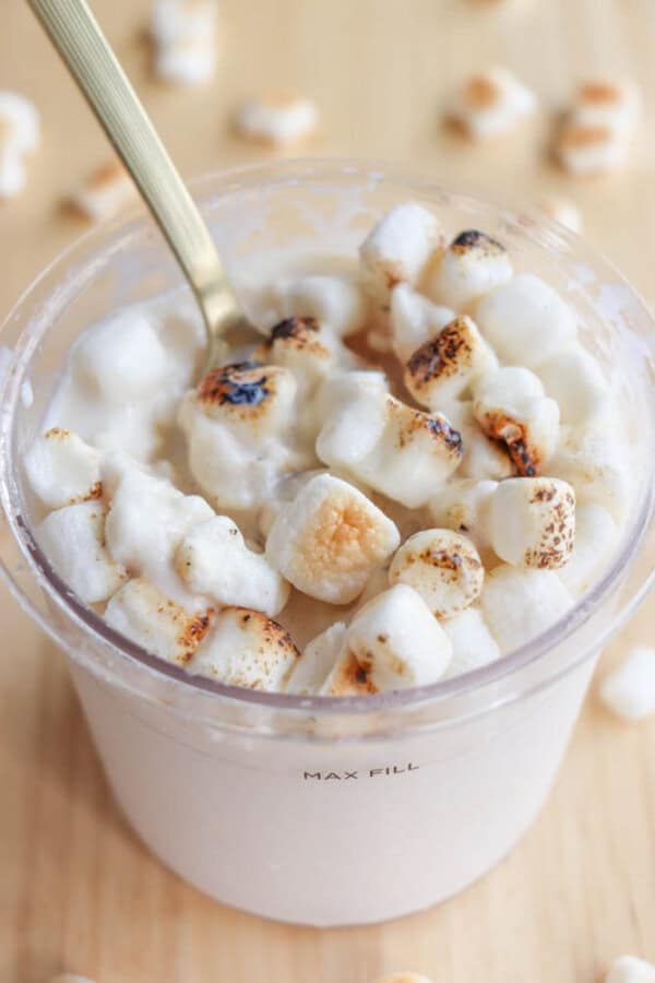 A plastic container filled with creamy yogurt topped with toasted mini marshmallows. A spoon is dipped into the yogurt, and a few marshmallows are scattered on a light wooden surface around the container.