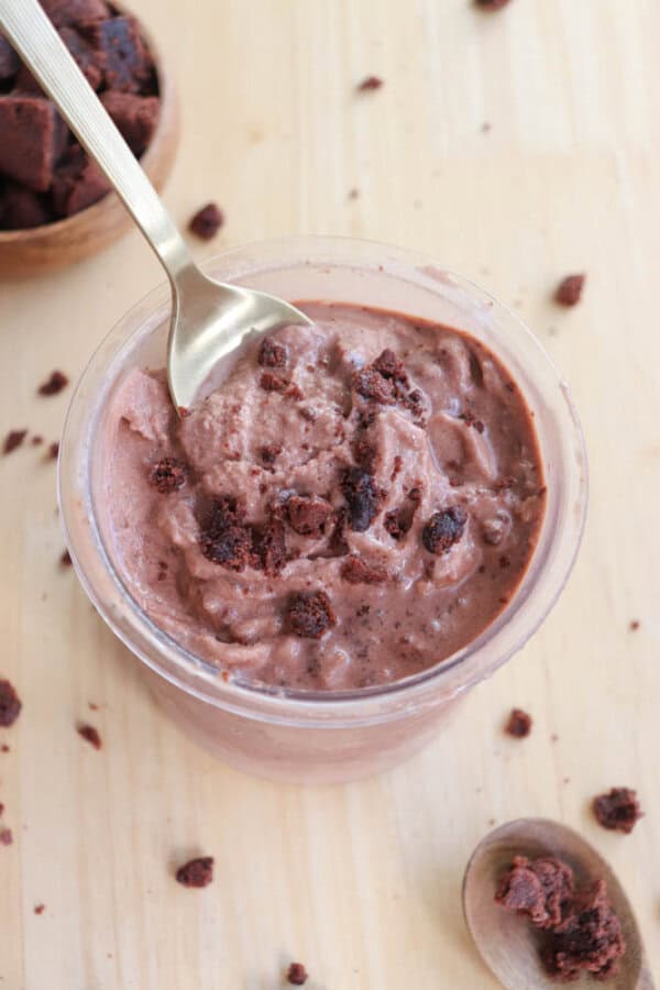 A pint with creamy chocolate ice cream topped with brownie chunks. A spoon is dipped into the ice cream, and more brownie pieces are scattered on the light wooden surface around the glass.