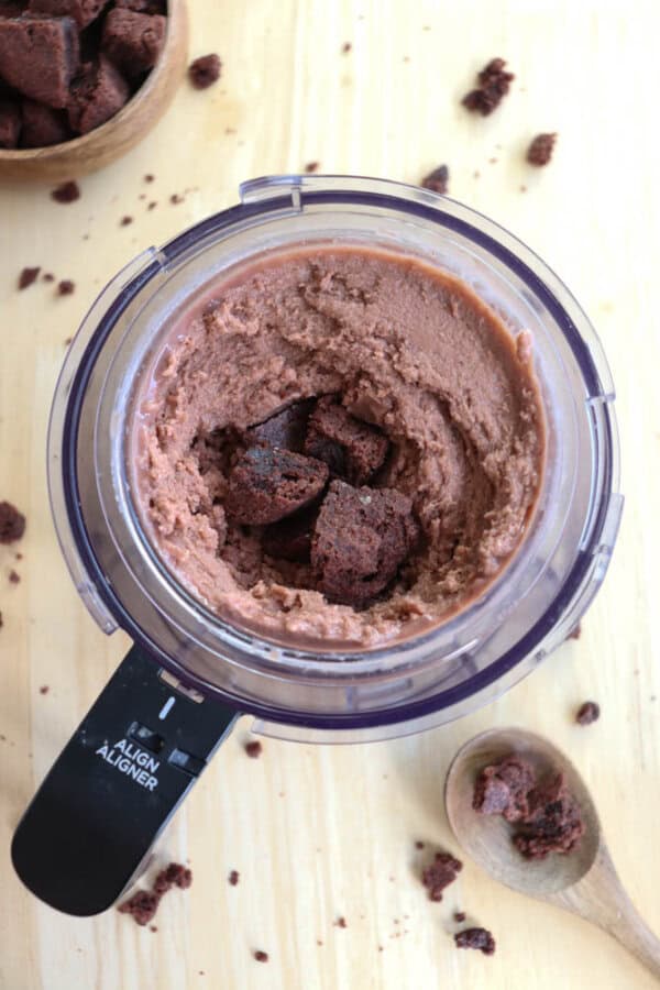A food processor with smooth brown cookie dough and chunks of cookies on top. A wooden spoon with cookie crumbs is beside it. A bowl of cookie pieces is partially visible in the corner. The scene is on a light wood surface.