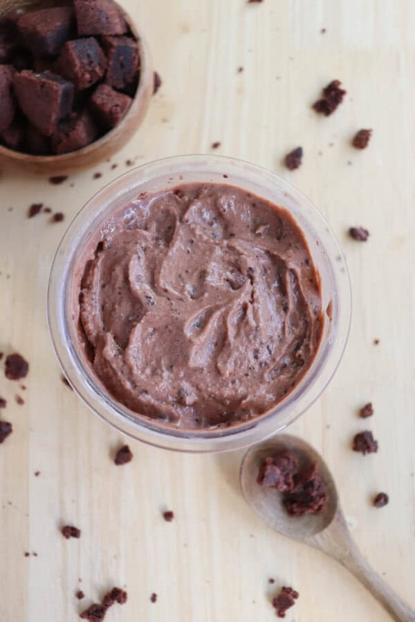 A bowl of chocolate ice cream with small chunks is centered on a wooden surface. Nearby, there are scattered chocolate chunks and a wooden spoon with more dough. A wooden bowl containing additional chocolate chunks is in the top left corner.
