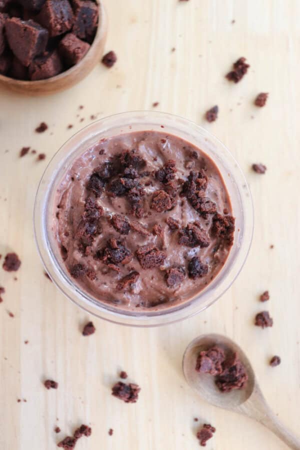 A glass bowl filled with chocolate ice cream topped with crumbled brownie pieces. A wooden spoon with brownie crumbs and a small wooden bowl with more brownies are on a light wooden surface. Brownie crumbs are scattered around.