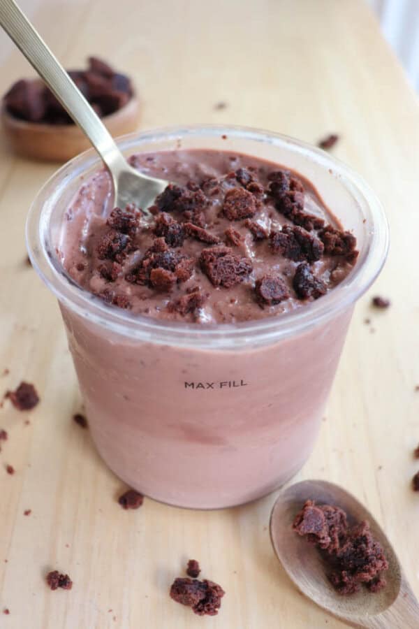 A plastic cup filled with chocolate ice cream, topped with crumbled brownie pieces. A spoon is inserted into the ice cream. A wooden table is visible, with scattered brownie crumbs and a wooden dish in the background.