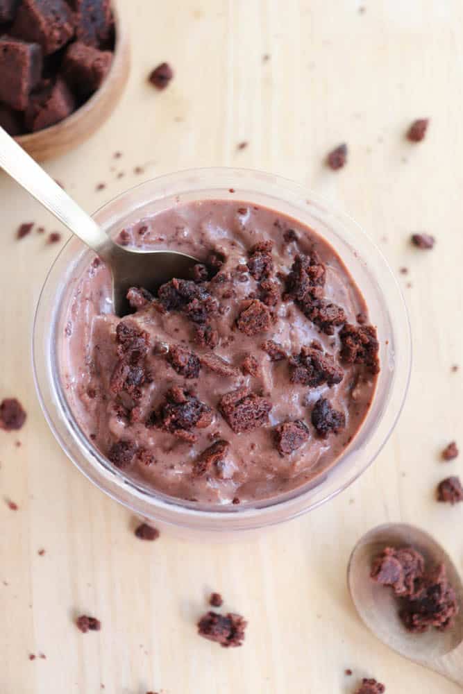 A glass jar filled with creamy chocolate pudding topped with chunks of brownie. A spoon is resting inside the jar. Small brownie pieces are scattered around on the wooden surface.