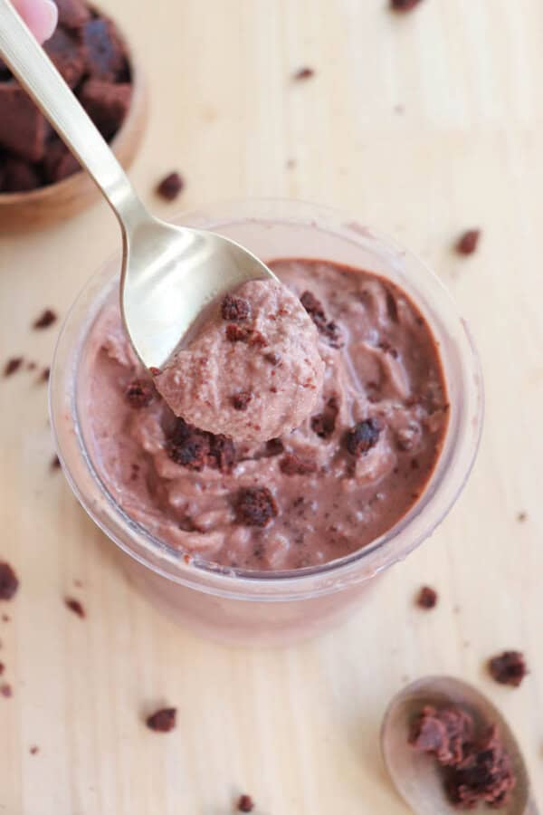 A spoonful of chocolate ice cream with brownie chunks is held above a container filled with the same dessert. The ice cream is creamy and chocolatey, and a wooden table surface is visible beneath. A bowl with more brownies is partially visible.
