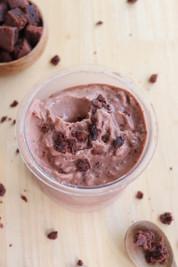 A clear cup filled with creamy chocolate mousse, topped with chocolate chunks. Several small chocolate pieces are scattered around the cup on a light wooden surface. A spoon and a small bowl with more chocolate chunks are in the background.