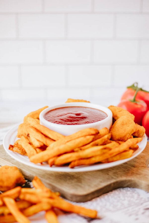 A plate of crispy fries and breaded chicken tenders surrounds a bowl of dipping sauce. Fresh tomatoes are in the background, all set on a wooden surface with a white tiled wall.