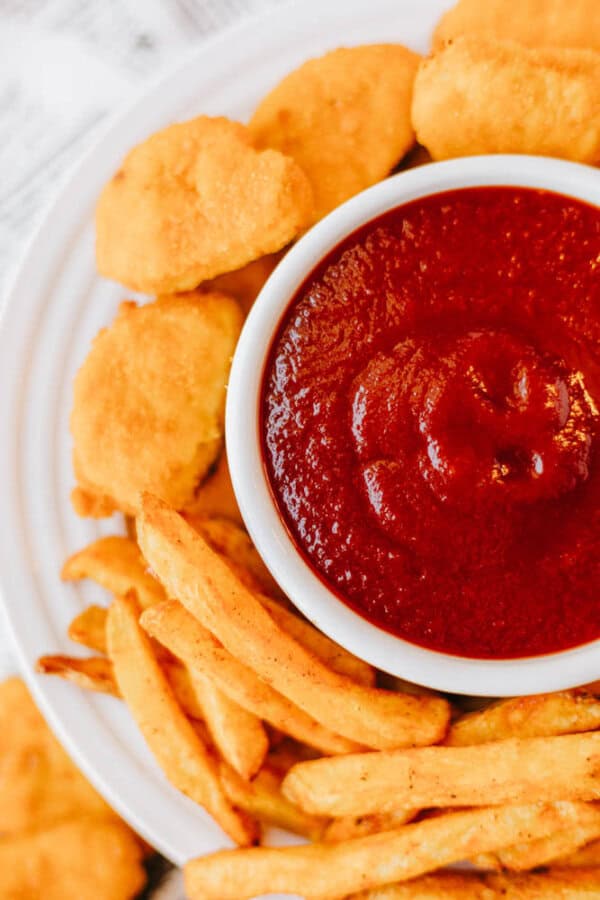 A plate of golden chicken nuggets and crispy fries surrounds a bowl of thick, dark red ketchup. The arrangement is on a white background, creating a contrast that highlights the vibrant colors of the meal.