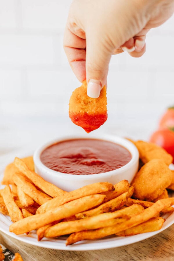 A hand holding a nugget dipped in a white bowl of red sauce. The bowl is on a plate with crispy fries and more nuggets. There are tomatoes in the blurred background on the wooden surface.