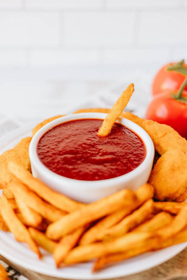 A plate of golden-brown fries surrounds a white bowl of ketchup. An onion ring rests on the rim, and a single fry is dipped into the sauce. Two ripe tomatoes are visible in the background.