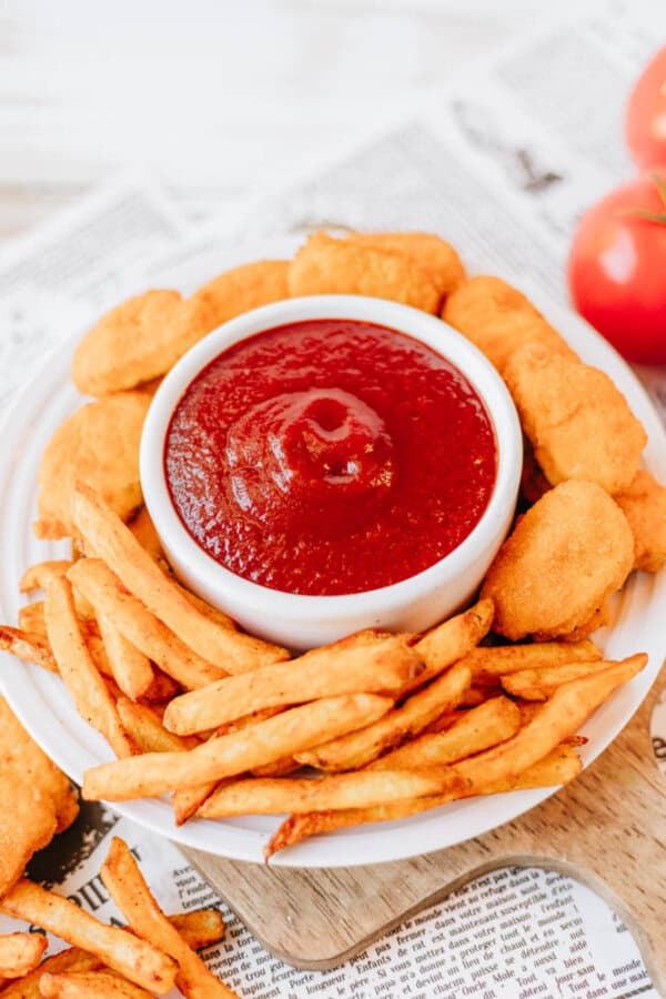 A white plate with crispy chicken nuggets and golden French fries surrounds a small bowl of red ketchup. The plate is placed on a newspaper, and there are two tomatoes partially visible in the background.