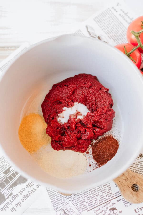 A white bowl contains a mound of tomato paste, garlic powder, onion powder, and chili powder. The ingredients are arranged separately. Fresh tomatoes and a wooden spoon are partially visible in the background.