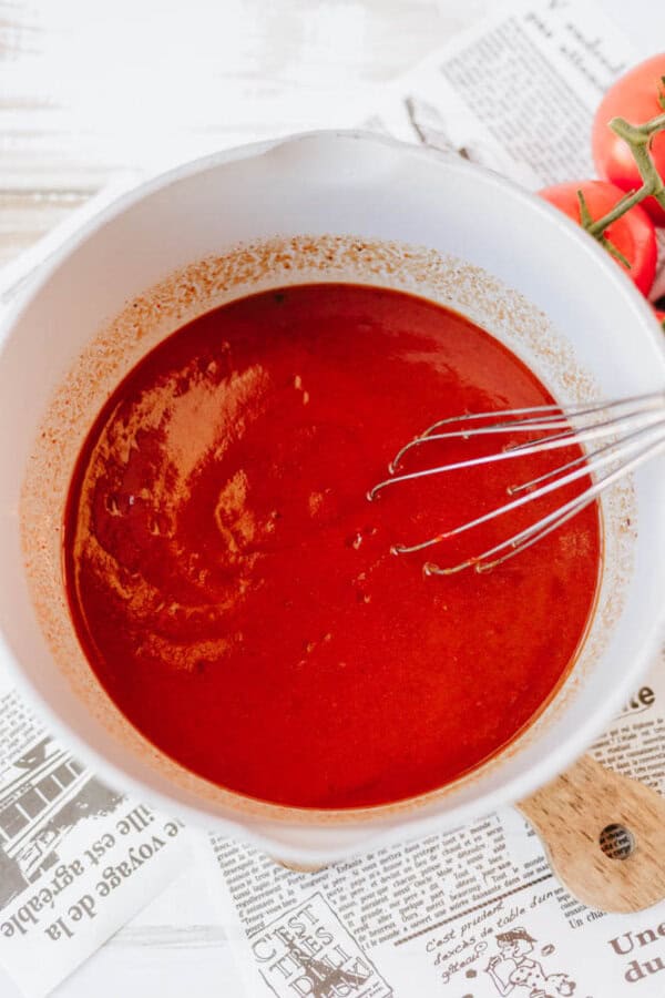 A bowl of rich red sauce with a whisk inside, placed on a table covered with newspaper. Fresh tomatoes are partially visible in the corner, suggesting the sauce is tomato-based.