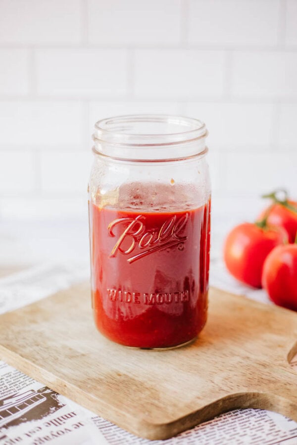 A glass jar labeled Ball filled with red tomato sauce sits on a wooden cutting board. Fresh tomatoes are in the background. The scene is set against a white tiled wall, with a newspaper partially visible.