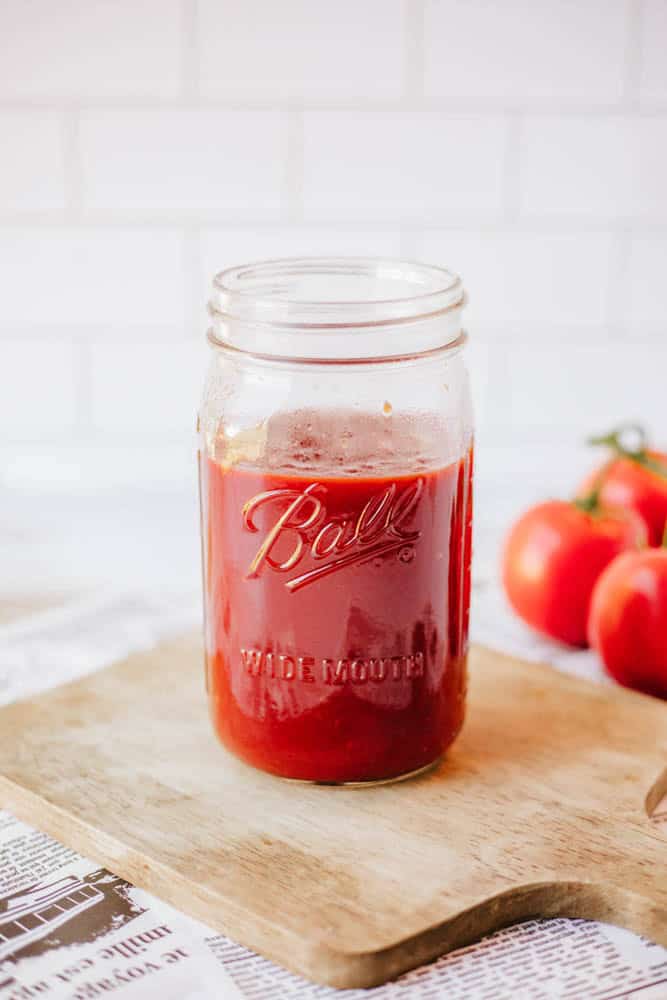 A glass jar labeled Ball filled with red tomato sauce sits on a wooden cutting board. Fresh tomatoes are in the background. The scene is set against a white tiled wall, with a newspaper partially visible.