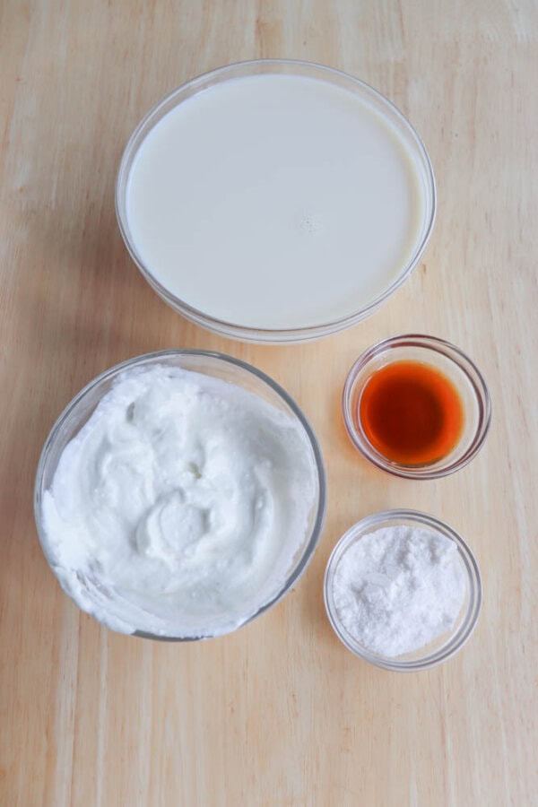 Four bowls on a wooden surface: large bowl with white liquid, medium bowl with whipped mixture, small bowl with white powder, and smallest bowl with amber liquid.