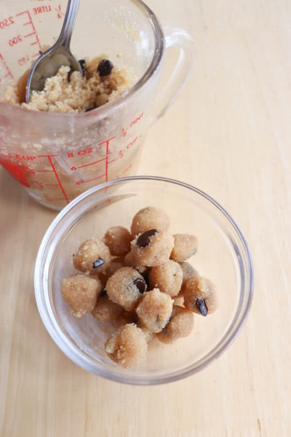 Glass bowl with round cookie dough bites featuring chocolate chips. Behind it, a measuring cup with remaining dough and a spoon. Wooden table surface.