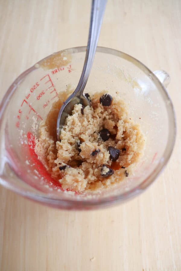 A glass measuring cup filled with cookie dough, containing chocolate chips, and a spoon sticking in it. The dough appears coarse and slightly crumbly, set on a light wooden surface.