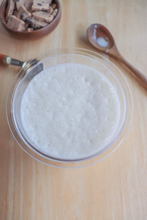 A frothy, white liquid in a transparent bowl on a wooden surface evokes the essence of high protein indulgence. Nearby, a wooden spoon with powder residue hints at the flavors within, while a small bowl filled with square beige chunks suggests cookie dough ice cream delights.