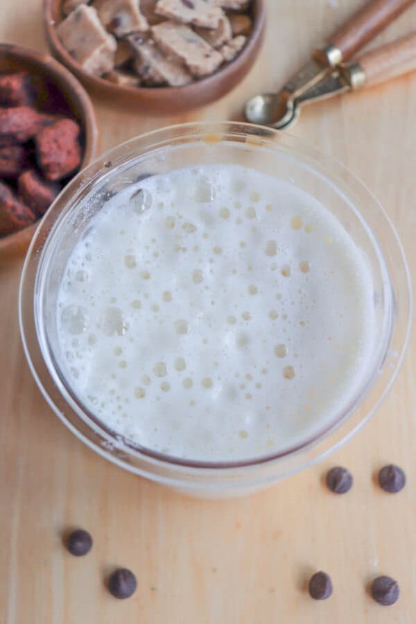A glass of frothy milk sits on a wooden surface. Around it are chocolate chips and bowls of chopped cookies and brownies. Three wooden spoons with brass handles are placed nearby.