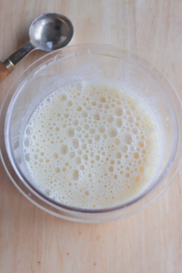 A clear bowl filled with frothy, creamy liquid reminiscent of a copycat Orange Julius recipe sits on a wooden surface. A metal measuring spoon with a wooden handle rests nearby.