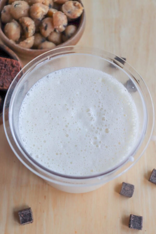 A glass filled with frothy milk is centered on a wooden surface. Surrounding the glass are small chocolate squares and a bowl of round cookies in the background.