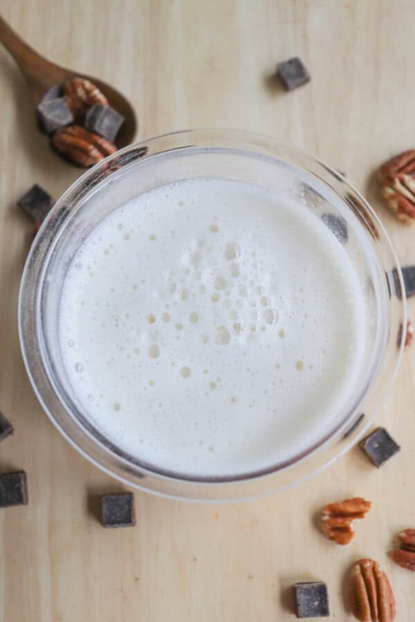 A top view of a pint of frothy protein shake on a wooden surface, surrounded by chocolate chunks and pecans. A wooden spoon holding more chocolate and pecans is visible in the top left corner.