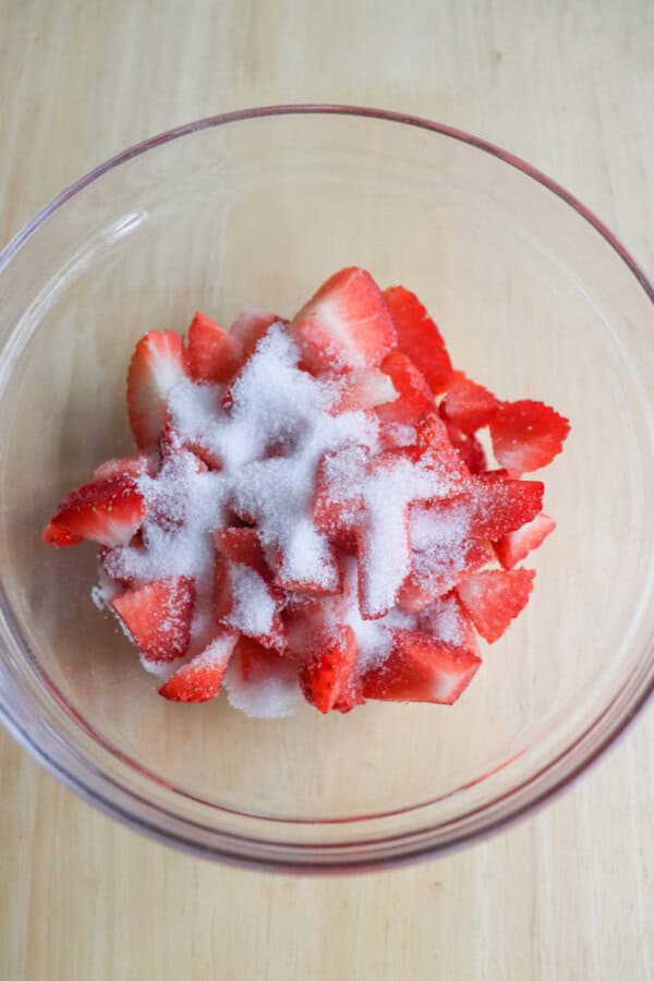 A clear glass bowl filled with sliced strawberries topped with a layer of white sugar, reminiscent of a raspberry cheesecake delight, placed on a light wooden surface.