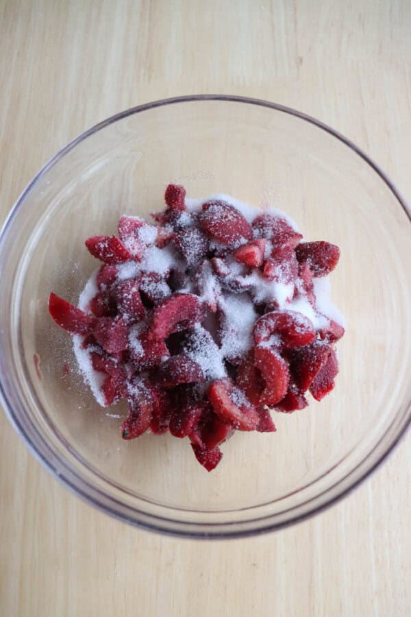 A clear glass bowl showcases sliced red cherries with a sprinkle of sugar, all displayed on a wooden surface.
