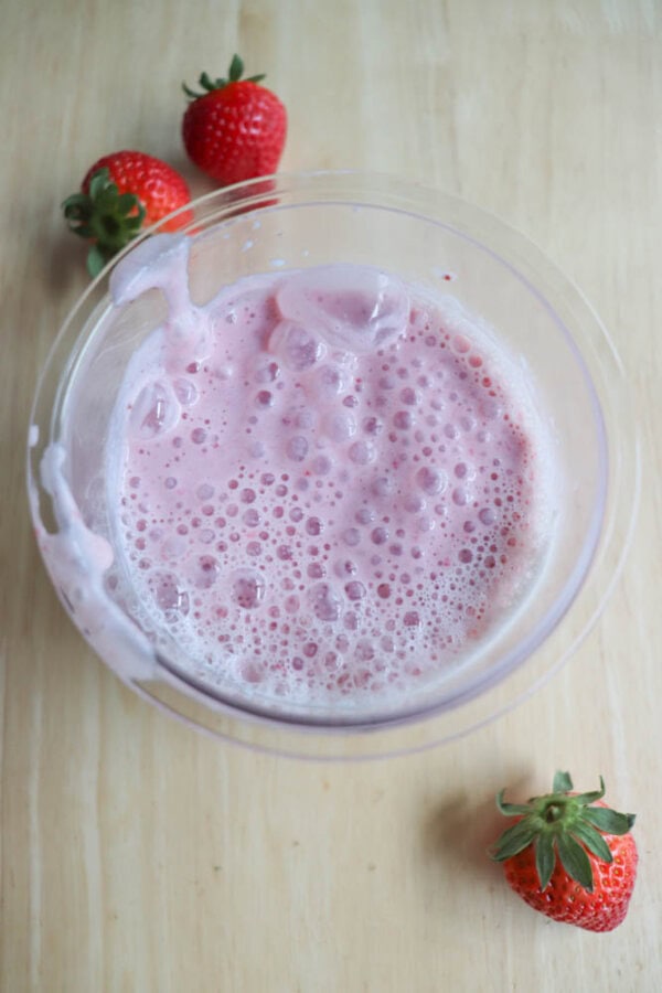 A plastic pint filled with a frothy pink smoothie sits on a wooden surface. Three whole strawberries are placed beside the bowl—two at the top left and one at the bottom right.