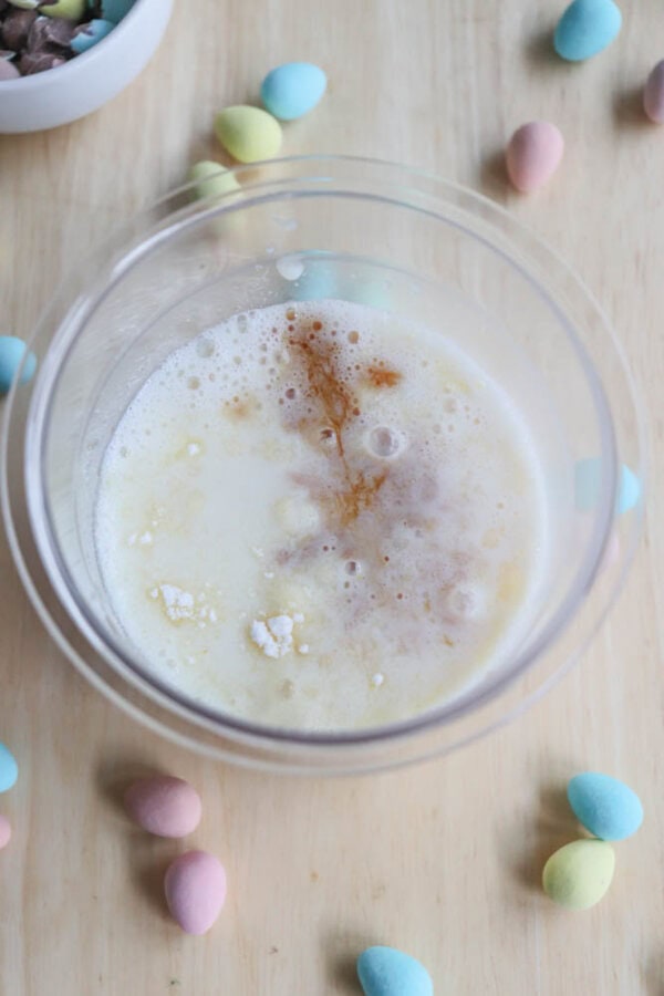 A glass cup filled with a creamy, frothy drink on a wooden surface is topped with cinnamon and powdered ingredients. Colorful pastel chocolate eggs resemble bunny tracks, playfully scattered around the cup.