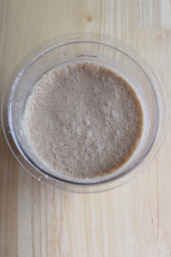 A top view of a clear container filled with a light brown liquid on a wooden surface. The mixture appears to be fermentation in progress, with a frothy texture on the surface.