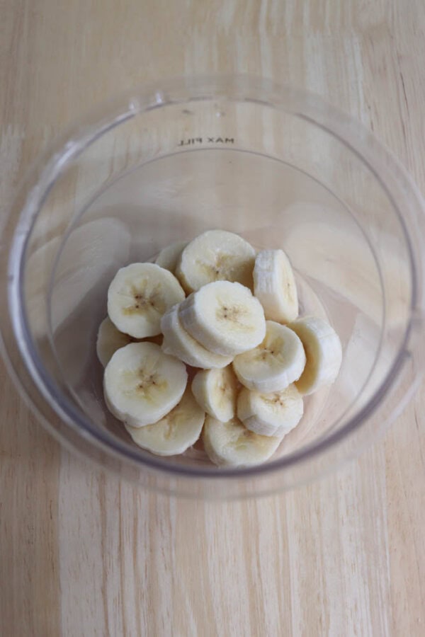 Sliced banana pieces in a clear glass blender jar on a wooden surface, viewed from above.