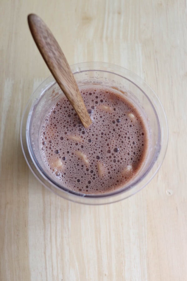 A glass of chocolate milk with small marshmallows floating on top, stirred with a wooden spoon, sits on a light wooden surface.