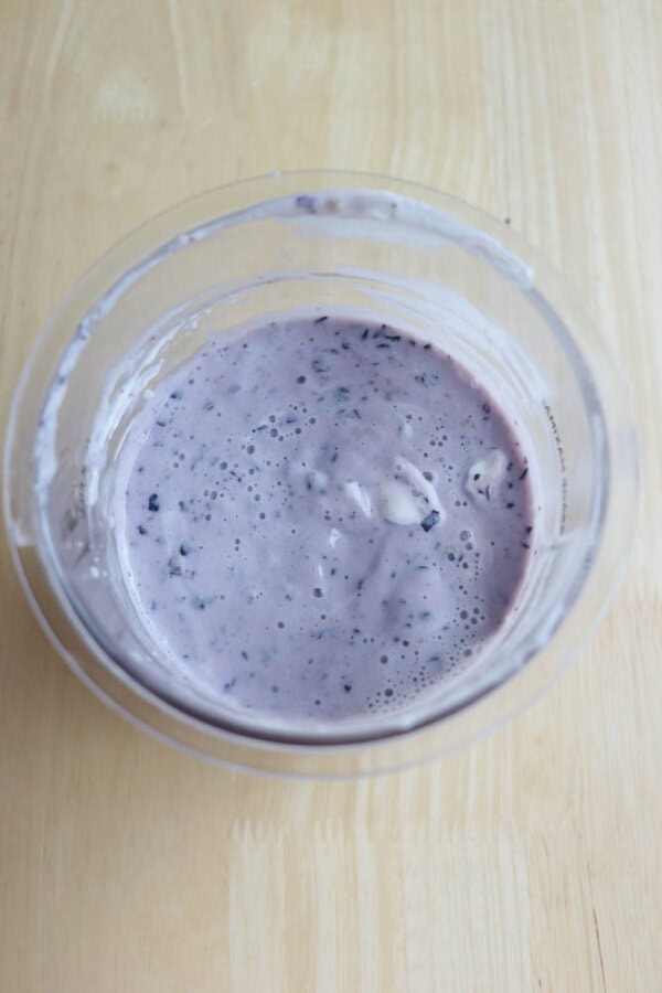 A clear plastic pint filled with a creamy, thick, purple mixture on a wooden surface. The mixture has small dark specks and a smooth texture, suggesting it could be a blueberry-flavored yogurt or smoothie.