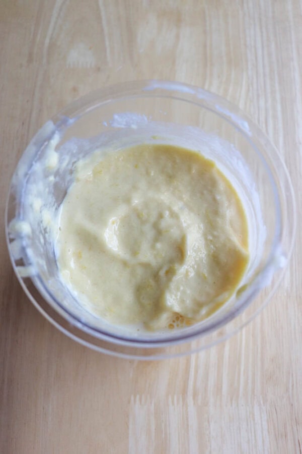 A clear plastic bowl filled with creamy, mango yogurt sits on a light wooden surface. The sauce appears smooth and thick, with some texture visible on the surface.
