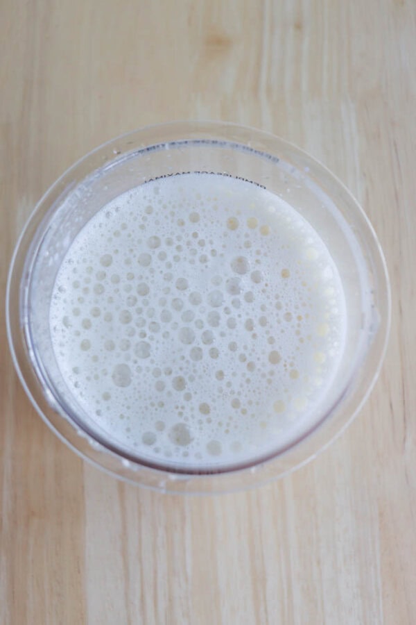 A clear plastic container filled with frothy, pale liquid on a light wooden surface. The liquid has a bubbly texture, resembling a milkshake or smoothie.