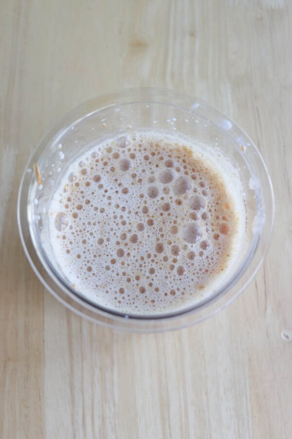 A pint of frothy peanut butter protein shake sits on a light wooden surface. The bubbles cover the top, indicating a freshly stirred or blended drink. The bowl is simple, with a clear rim, allowing a view of the contents.