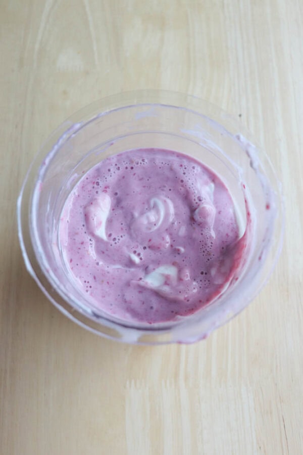 A glass bowl filled with pink yogurt is placed on a light wooden surface. The yogurt appears creamy with visible swirls and flecks of darker pink.