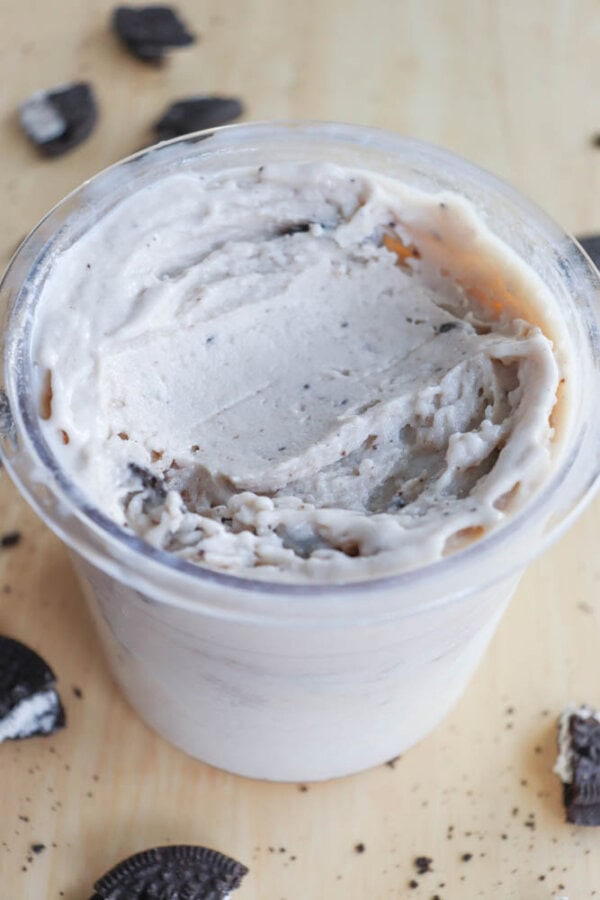 A clear plastic cup brimming with creamy blueberry cheesecake ice cream mixed with crushed cookies rests on a wooden surface. Broken cookie pieces are scattered around, enhancing the visual indulgence. The ice cream appears smooth and slightly swirled, inviting a delectable experience.