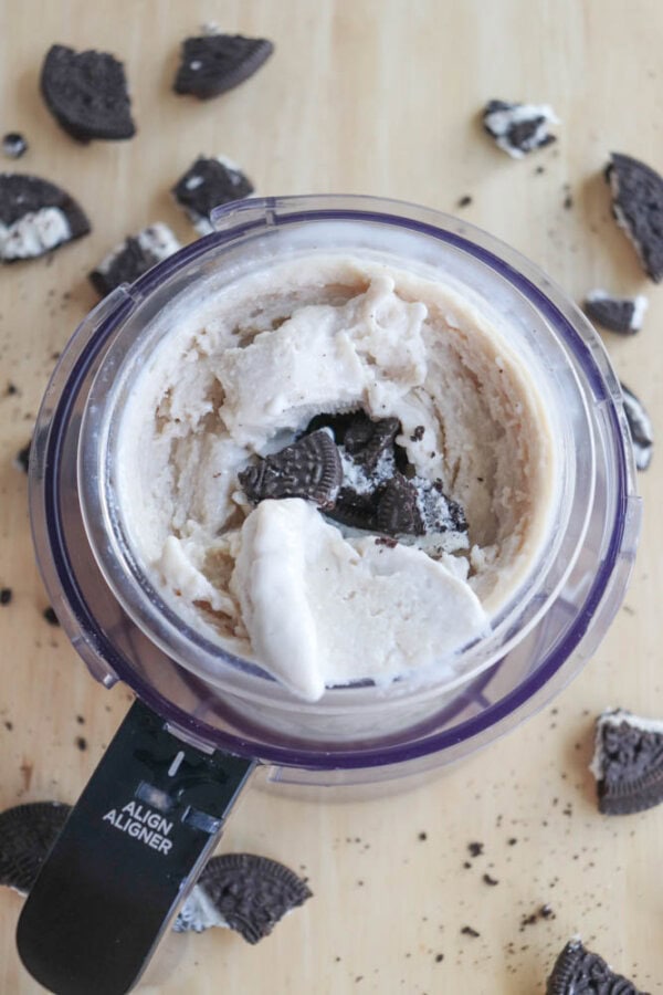In a food processor, blueberry cheesecake ice cream mixture is blended with crushed chocolate sandwich cookies. Additional pieces of these cookies are scattered around the processor on a wooden surface.