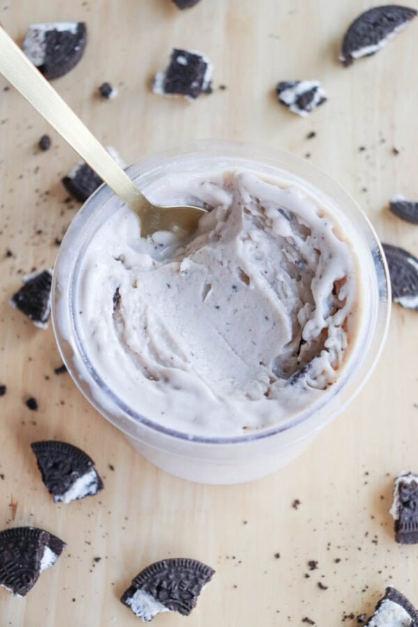 A container of cookies and cream ice cream with a gold spoon scooping into it. Broken chocolate cookie pieces with cream filling are scattered around on a light wooden surface, creating a dreamy contrast reminiscent of blueberry cheesecake ice cream dreams.