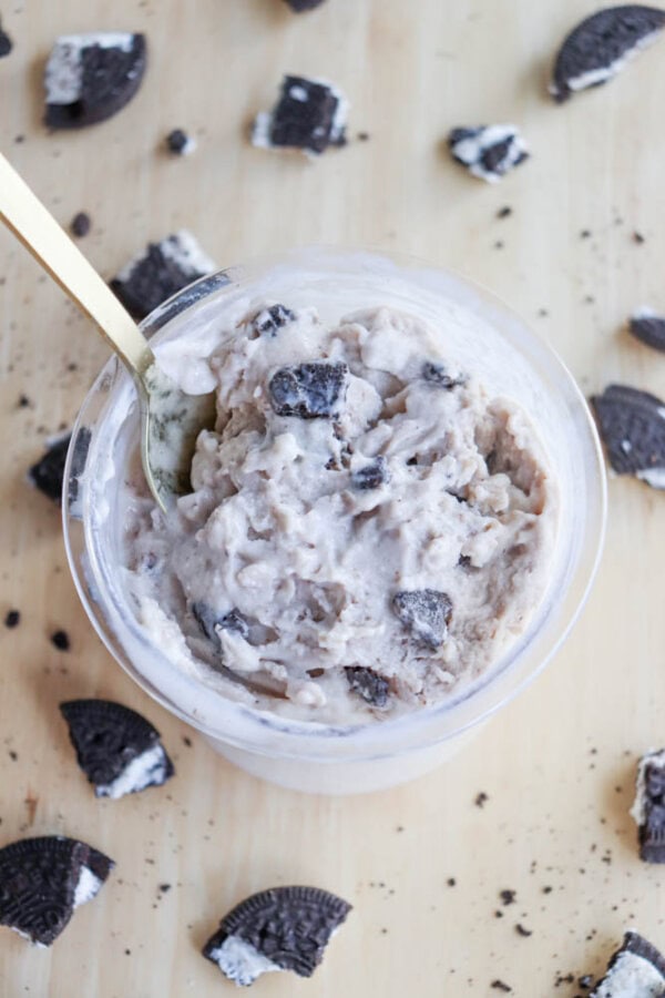 A creamy dessert with a cookies and cream flavor, reminiscent of blueberry cheesecake ice cream, is presented in a round, clear container on a wooden surface. A gold spoon dips into the dessert, surrounded by scattered broken cookies.