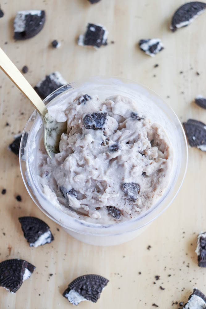 A creamy dessert with a cookies and cream flavor, reminiscent of blueberry cheesecake ice cream, is presented in a round, clear container on a wooden surface. A gold spoon dips into the dessert, surrounded by scattered broken cookies.