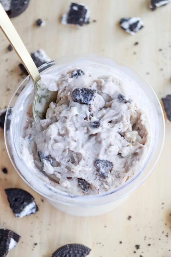 A cup of Oreo ice cream with chunks of cookies, served with a gold spoon on a light wooden surface. Nearby, hints of blueberry cheesecake ice cream add a fruity contrast, while Oreo pieces are scattered around the cup to highlight the creamy texture and cookie bits within.