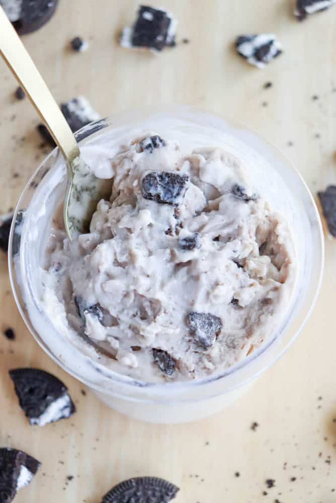 A cup of Oreo ice cream with chunks of cookies, served with a gold spoon on a light wooden surface. Nearby, hints of blueberry cheesecake ice cream add a fruity contrast, while Oreo pieces are scattered around the cup to highlight the creamy texture and cookie bits within.
