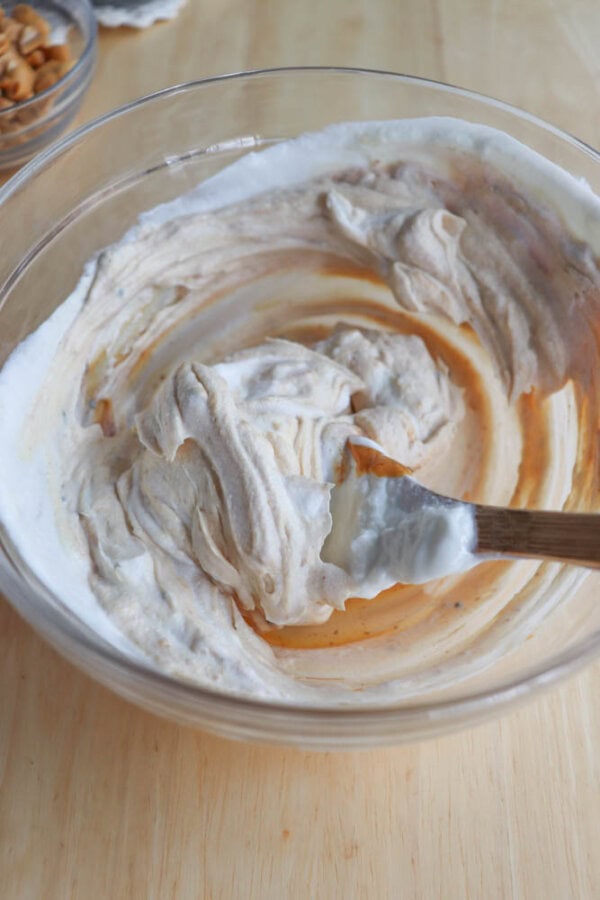 A glass bowl with a creamy mixture being stirred with a wooden spoon. The mixture appears to be a blend of white and light brown swirls, indicating a possible mixing of different ingredients. The bowl is on a wooden surface.